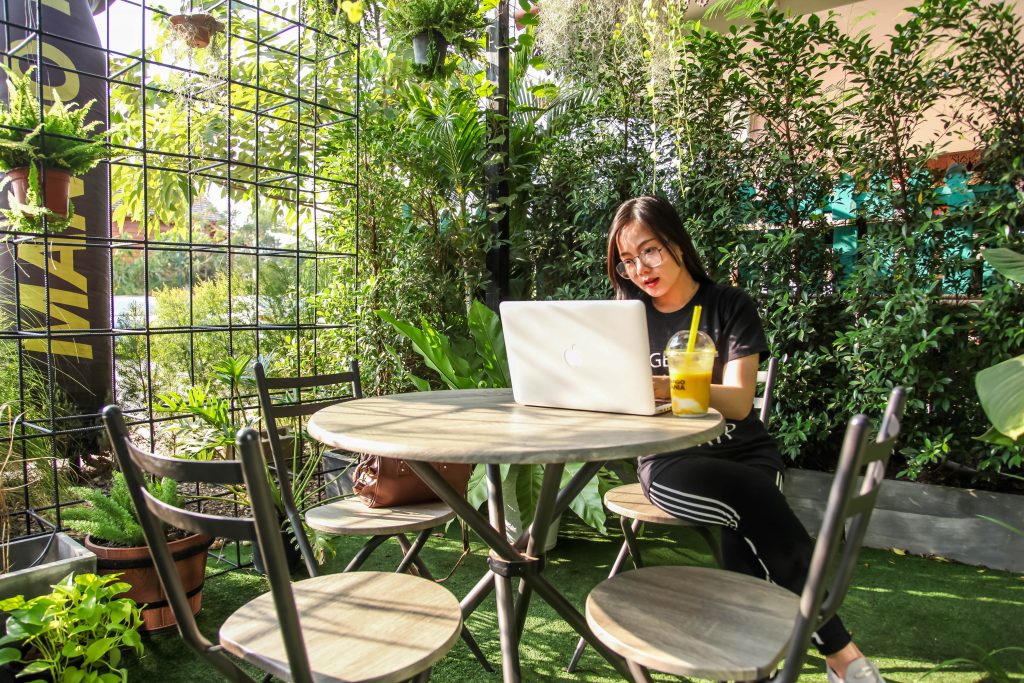 Young woman sitting at outdoor cafe seating working on laptop with drink beside her
