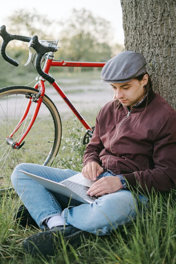 man working under tree on laptop wearing windbreaker and cap with bike in background